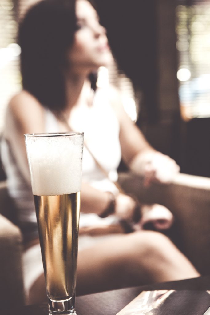Tall glass of beer with frothy head on a table, blurred background of a person sitting in a cozy setting